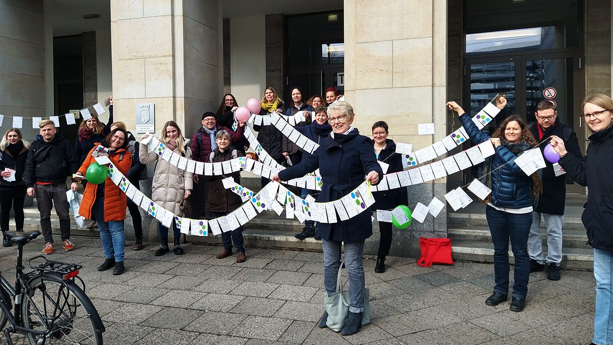 Kita-Sozialarbeiter:innen aus ganz Berlin stehen vor der Senatsverwaltung für Bildung und halten Schnüre mit Postkarten hoch