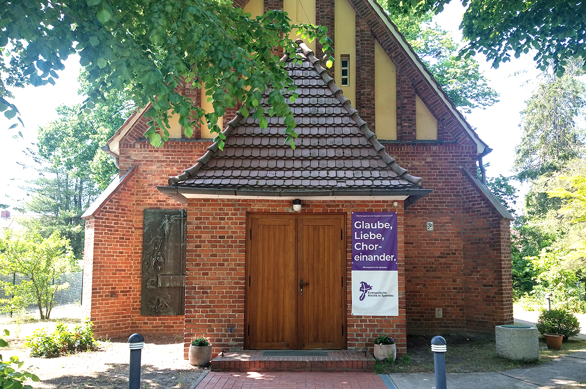 die kleine Kirche Staaken-Gartenstadt, umgeben von Bäumen, neben dem Eingang ein Banner "Glaube, Liebe, Choreinander"