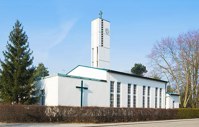 Gnadenkirche, modernes weißes Gebäude, über dem Hauptgebäude ragt ein Turm in die Höhe, an einer Wand ist ein große Kreuz angebracht