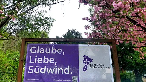 Banner "Glaube, Liebe, Südwind" vor blühenden Bäumen