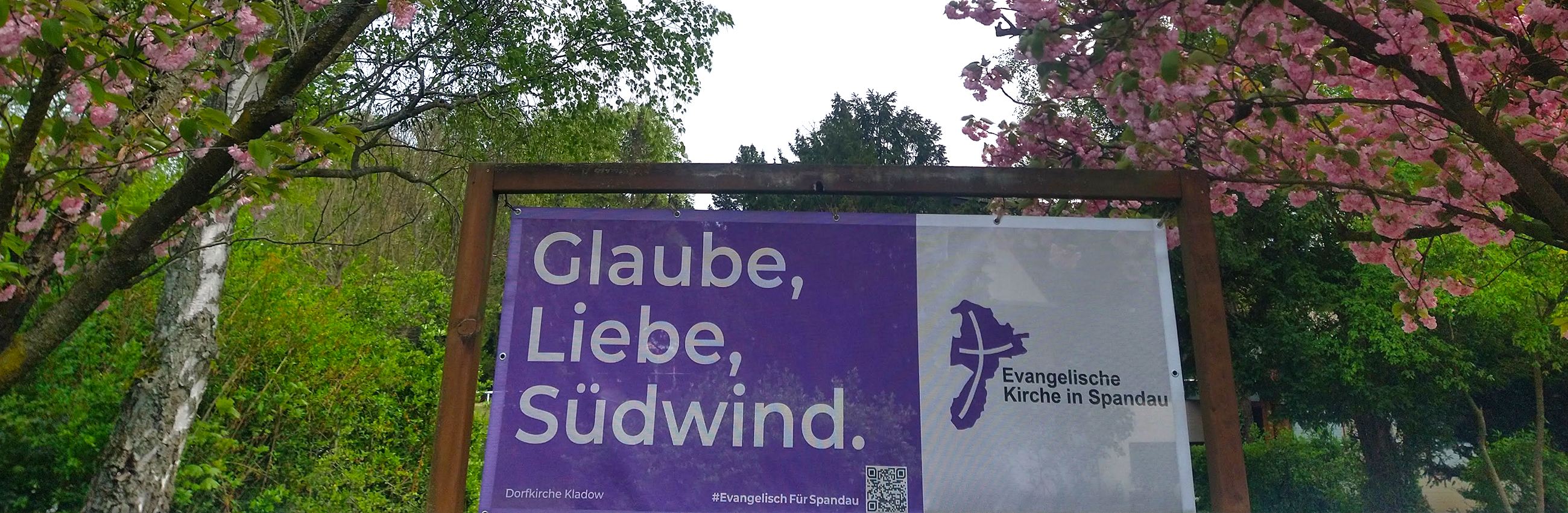 Banner "Glaube, Liebe, Südwind" vor blühenden Bäumen