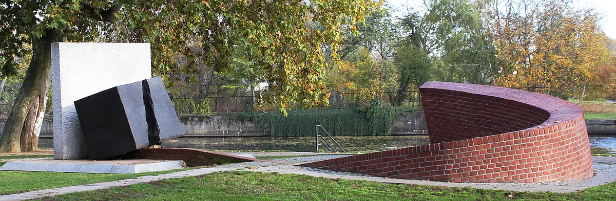 Das Mahnmal am Lindenufer: Ein dunkler Kubus mit Riss steht leicht angekippt vor einem hellen, schmalen, hohen Quader. Gegenüber steht eine gemauerte Spirale.