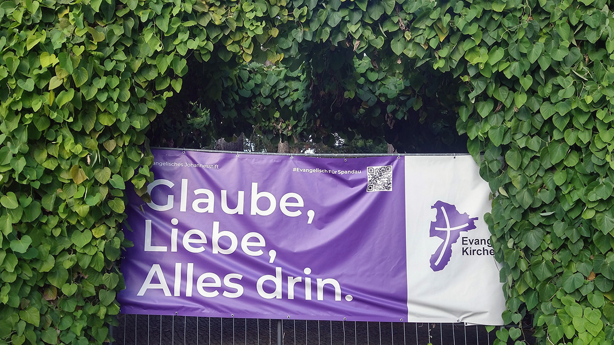Lila Banner "Glaube, Liebe, Alles drin.", das von grünen Rankpflanzen umgeben ist.