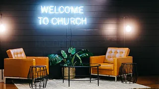 Symbolbild: Unter der Leuchtschrift "Welcome to Church" stehen zwei Sessel und ein Tisch - Wohnzimmerfeeling