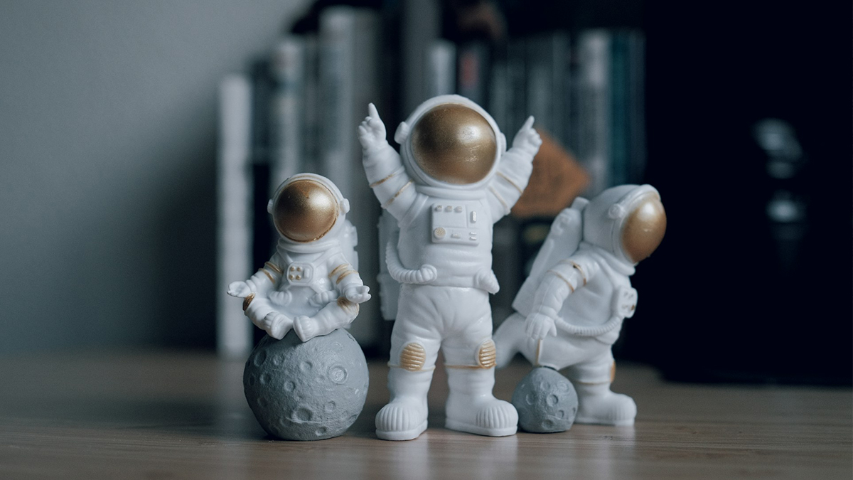 3 kleine Astronautenfiguren: der 1. sitzt auf einer Mondkugel, der 2. streckt die Arme triumphierend nach oben, der 3. spielt mit einer Mondkugel Fußball