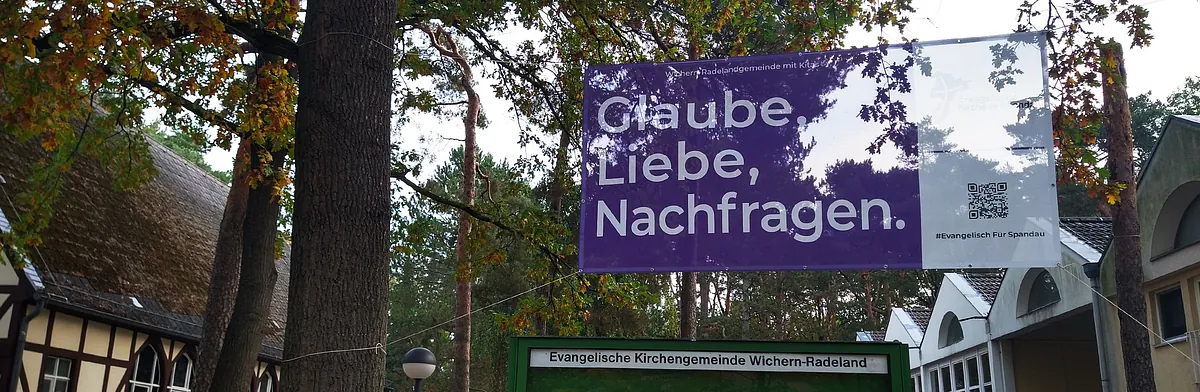 Zwischen Bäumen ist ein Banner aufgespannt "Glaube, Liebe, Nachfragen." links ist ein Fachwerkgebäude (Wichernkirche), rechts das moderne Gemeindegebäude