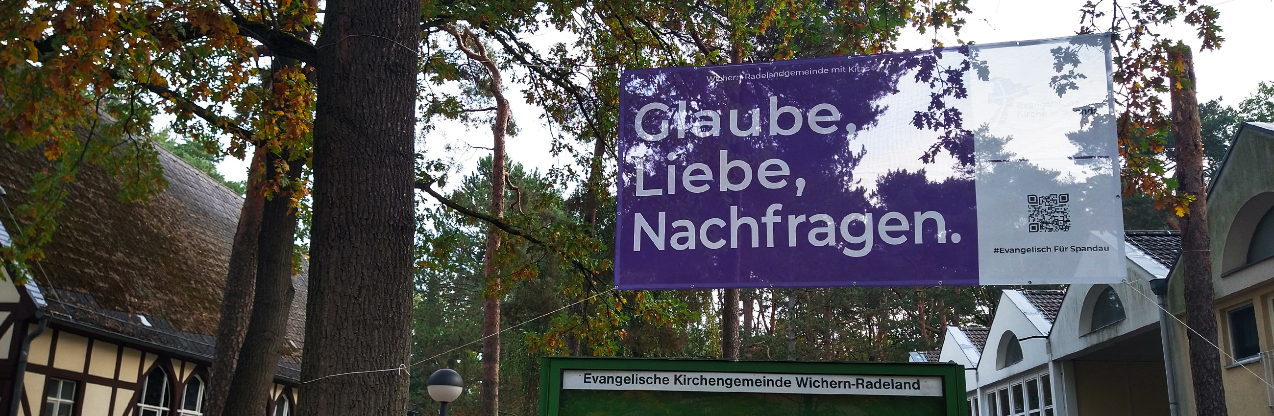 Zwischen Bäumen ist ein Banner aufgespannt "Glaube, Liebe, Nachfragen." links ist ein Fachwerkgebäude (Wichernkirche), rechts das moderne Gemeindegebäude