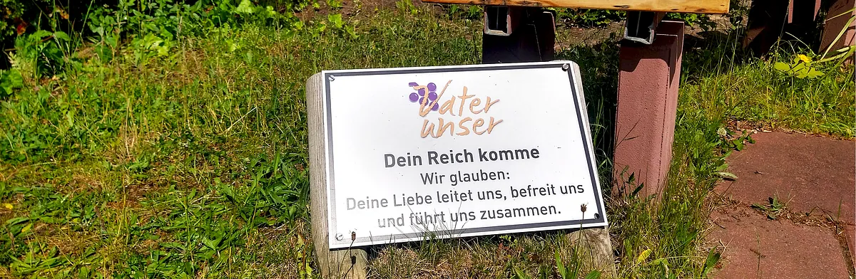 Eine Tafel im Gras: "Dein Reich komme / Wir glauben: Deine Liebe leitet uns, befreit uns und führt uns zusammen."