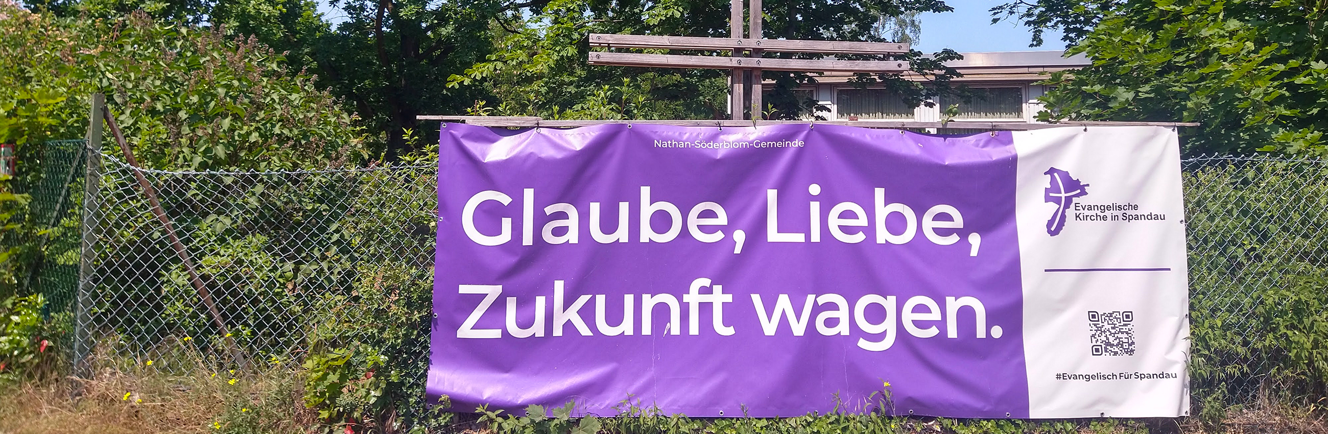 Ein lila Banner "Glaube, Liebe, Zukunft wagen" hängt an einem Drahtzaun, dahinter steht ein Holzkreuz.